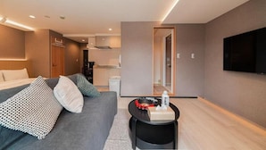 A modern hotel room with a grey sofa, a small black side table, a flat-screen TV mounted on the wall, and a visible kitchen area in the background. - SAIEI HOTEL (Tokyo)