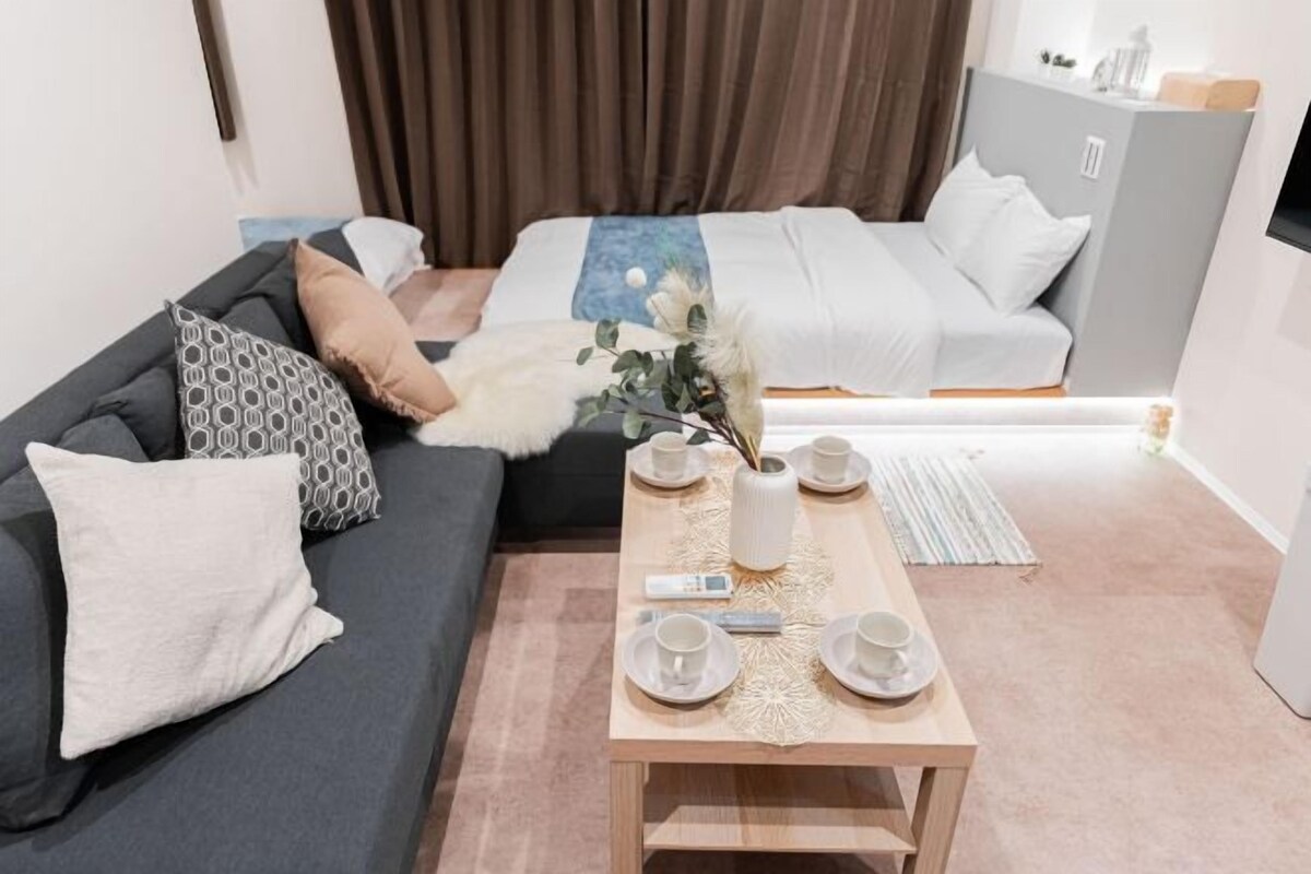 A modern hotel room with a grey sofa, a wooden coffee table with a vase and cups, and a bed with white bedding.
