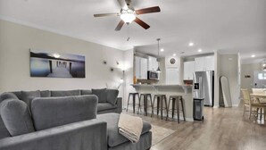 Un salon moderne avec un canapé gris, un ventilateur de plafond et un coin cuisine équipé d’un four à micro-ondes, d’un réfrigérateur et de tabourets de bar.
