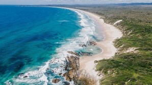 An aerial view of a coastal landscape with a sandy beach, rocky shoreline, and dense green vegetation. - 65 Ocean Roads, Brooms Head (Brooms Head)