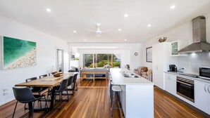A modern kitchen with a central island, wooden flooring, and a view of the outdoors through large windows. - 65 Ocean Roads, Brooms Head (Brooms Head)