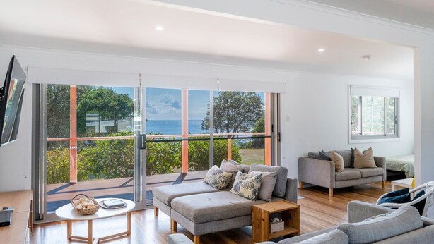 A spacious living area with a large sliding glass door leading to a balcony with a view of the sea. There are two sofas and a coffee table. - 65 Ocean Roads, Brooms Head (Brooms Head)