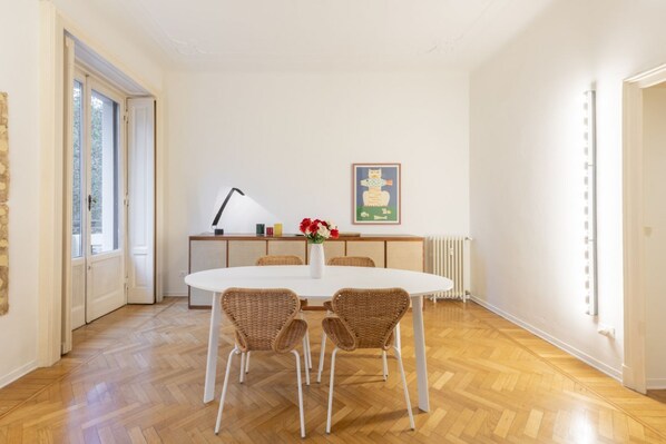 A room with a round table, four wicker chairs, a white lamp, a radiator, and a framed picture on the wall.