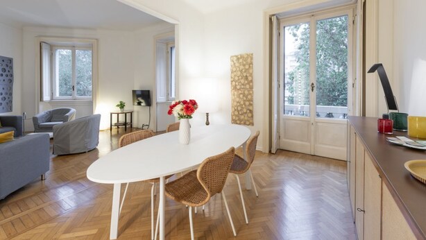 A dining area with a white table, wicker chairs, a vase with flowers, and a view through a glass door.