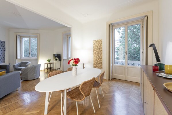 A dining area with a white table, wicker chairs, a vase with flowers, and a view through a glass door.