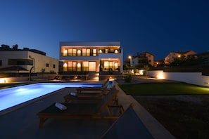 A modern two-story house with a swimming pool, lounge chairs, and a well-lit exterior at dusk.