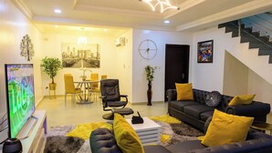 Una sala moderna con sofá negro, cojines amarillos, televisor de pantalla plana y comedor con mesa y sillas.