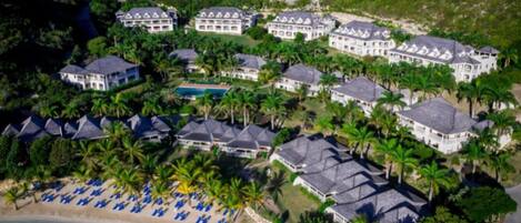 A resort with multiple buildings, a beach with blue umbrellas, and a pool.
