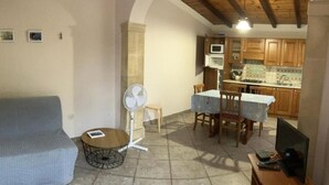 A living room with a sofa, a fan, a table, and a TV. There is a kitchen in the background with wooden cabinets and a tiled backsplash. - Apartment 1-Villa Augusta del Sol (Fontane Bianche)