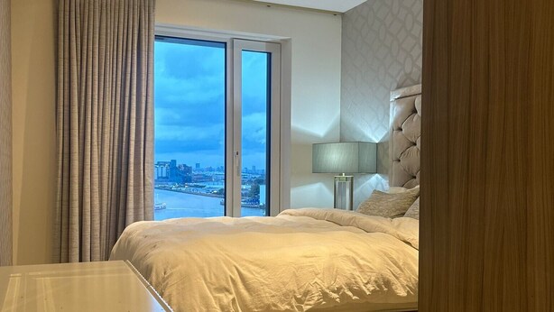 A hotel room with a bed, a desk, a chair, a lamp, and a view of the city through the window. - Cozy Riverview London Apartment Near the o2 Arena (London)