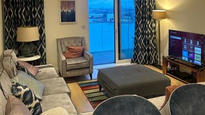 A modern living room with a sofa, armchairs, a coffee table, and a TV. There is a balcony with a view of the city. - Cozy Riverview London Apartment Near the o2 Arena (London)