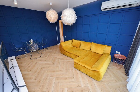 Un salon moderne avec un canapé jaune, deux luminaires sphériques suspendus, un parquet en bois disposé en chevrons et un mur bleu à panneaux.
