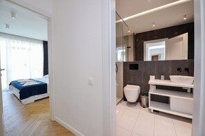 Une chambre d’hôtel moderne, équipée d’un lit, d’une salle de bain avec un lavabo et un miroir blancs, et d’une poubelle visible.