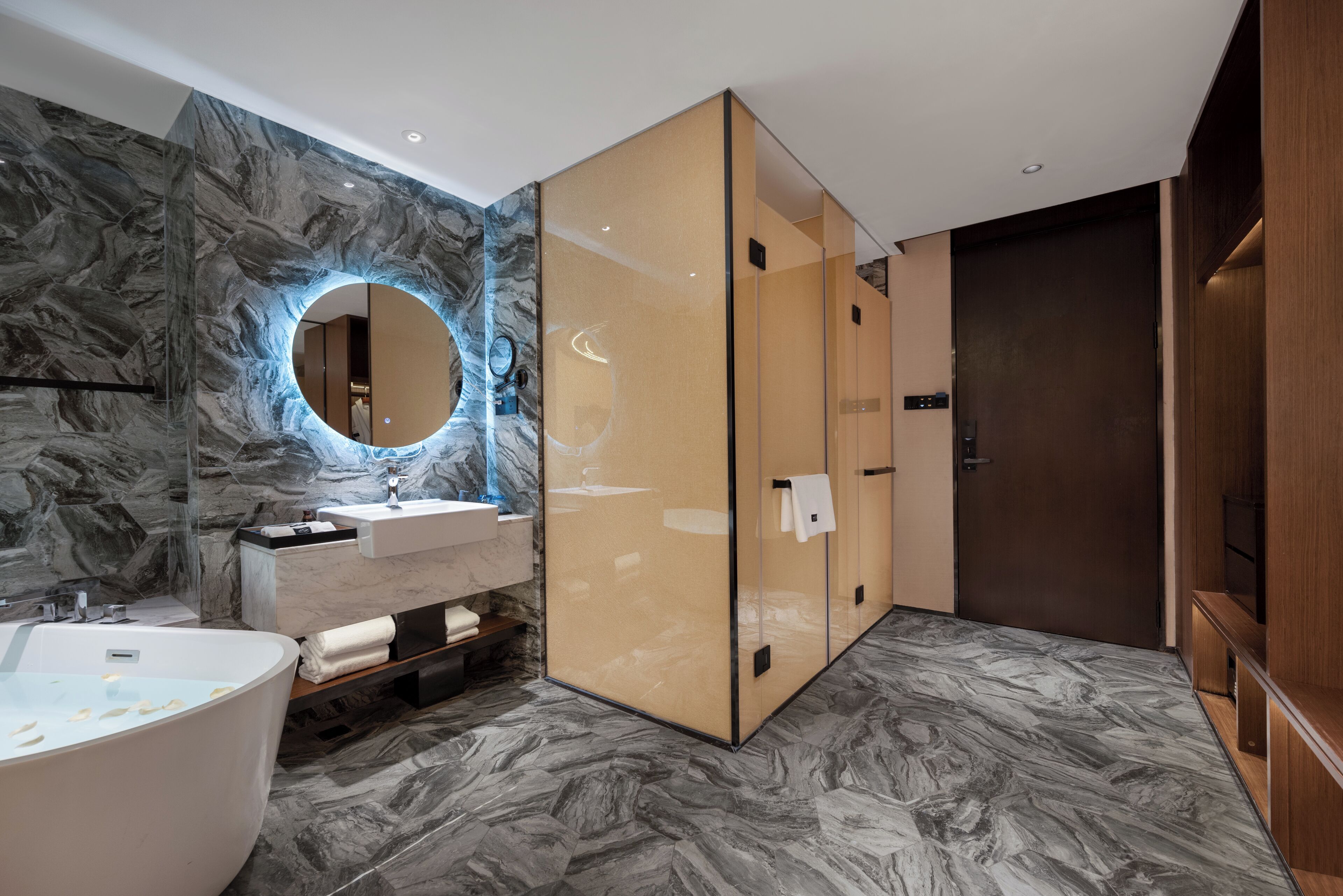 A modern bathroom with a marble wall, a round mirror, a freestanding bathtub, and a wooden vanity.
