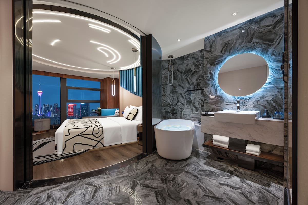Een moderne hotelkamer met een gebogen plafond, een groot bed, een badkuip en een badkamer met een marmeren wand en een ronde spiegel.