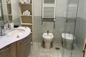 Une salle de bain moderne avec un lavabo rond, un bidet et des toilettes. On y trouve des étagères avec des serviettes et une cabine de douche en verre.