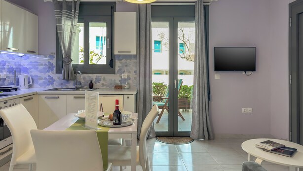 Une cuisine moderne avec des meubles blancs, un coin repas comprenant une table et des chaises, un téléviseur fixé au mur et une porte-fenêtre coulissante donnant sur un espace extérieur.