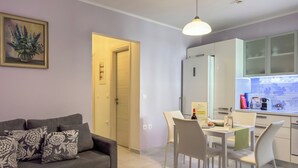 Un salon moderne avec un canapé gris, des chaises blanches et une table à manger. On aperçoit une cuisine avec des meubles blancs et un dosseret carrelé.