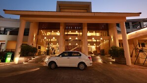 Entrada moderna de un hotel con un coche blanco estacionado frente a él, grandes columnas y un corredor cubierto.