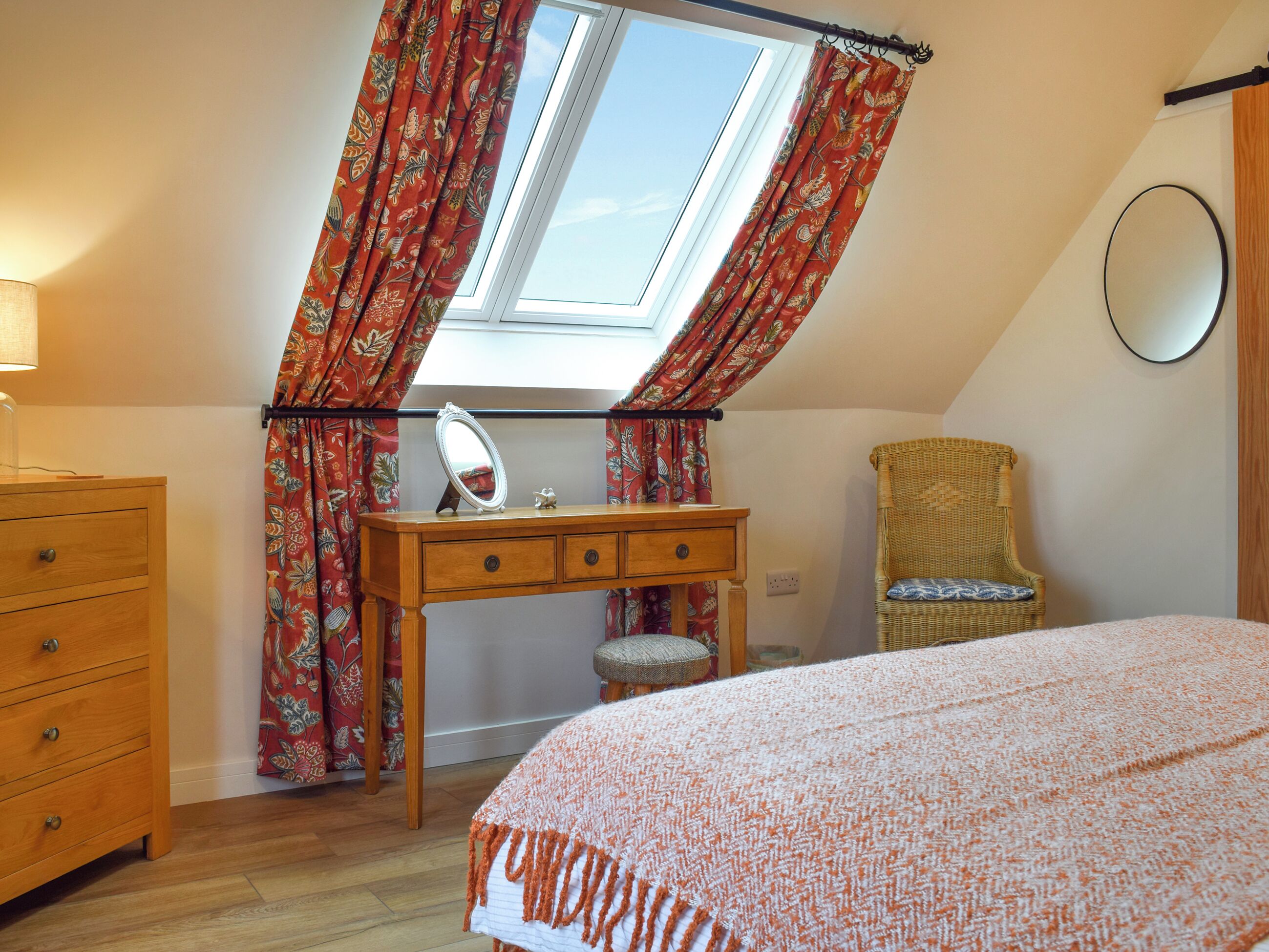 Een slaapkamer met een houten dressoir, een klein krukje, een rieten stoel, een bed met een beddegoed met patroon, een spiegel op het dressoir en een dakraam met gordijnen.