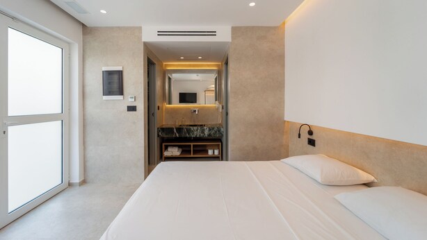 Una habitación de hotel moderna con una cama grande, un banco empotrado con espacio de almacenamiento, un espejo y un baño con encimera de mármol.