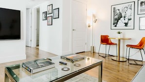 Een moderne woonkamer met een flatscreen-tv, een glazen salontafel met boeken en een afstandsbediening, twee oranje fauteuils rond een kleine tafel, en ingelijste foto’s aan de muur.