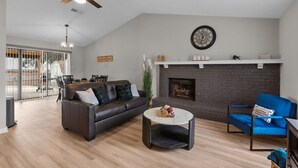 A living room with a fireplace, a ceiling fan, and a dining area with a glass door leading outside. - DFW Retreat • Pool, Spa & Minutes to Dallas & Fort Worth (Flower Mound)