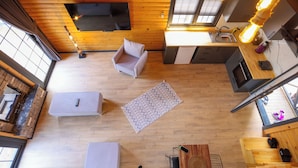 A compact living space with a wooden floor, a sofa, a small table, a kitchen area with a fridge, and a television mounted on the wall.