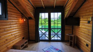 A log cabin room with a bed, a bench, a TV, and a view of a green landscape through a large window.