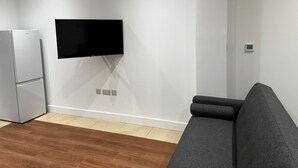 A modern living room with a flat-screen TV mounted on the wall, a grey sofa, and a stainless steel refrigerator.