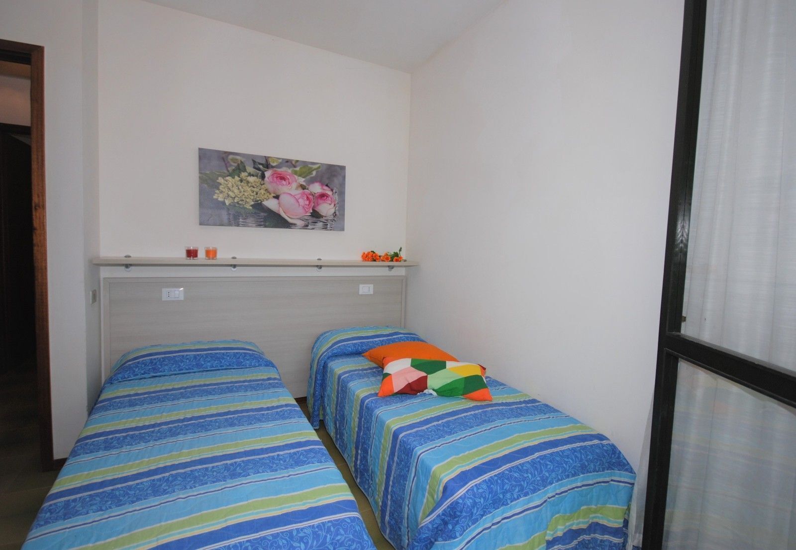 A small room with two beds, a painting on the wall, and a window with curtains.