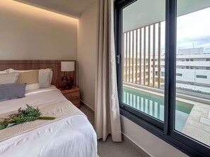 A hotel room with a large window, a bed with white linens, a nightstand with a lamp, and a view of a building complex.