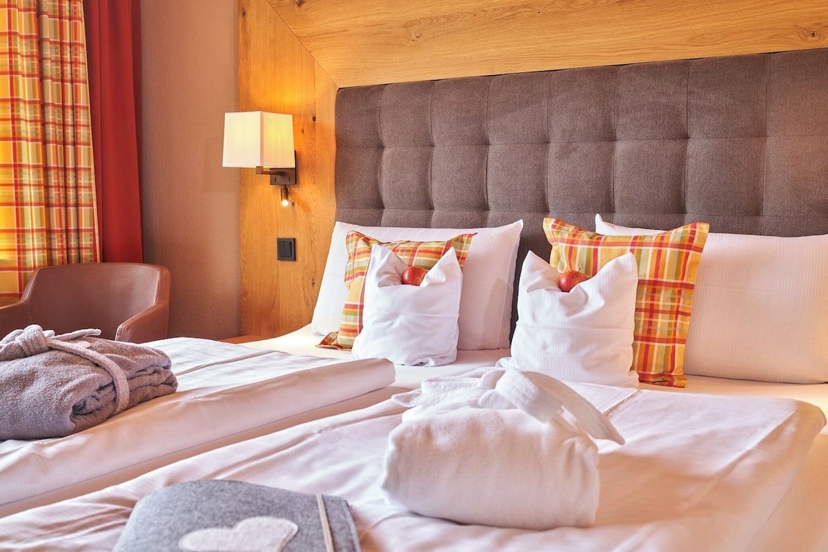 A hotel room with a tufted headboard, white bedding, plaid pillows, and a lamp on the wall.