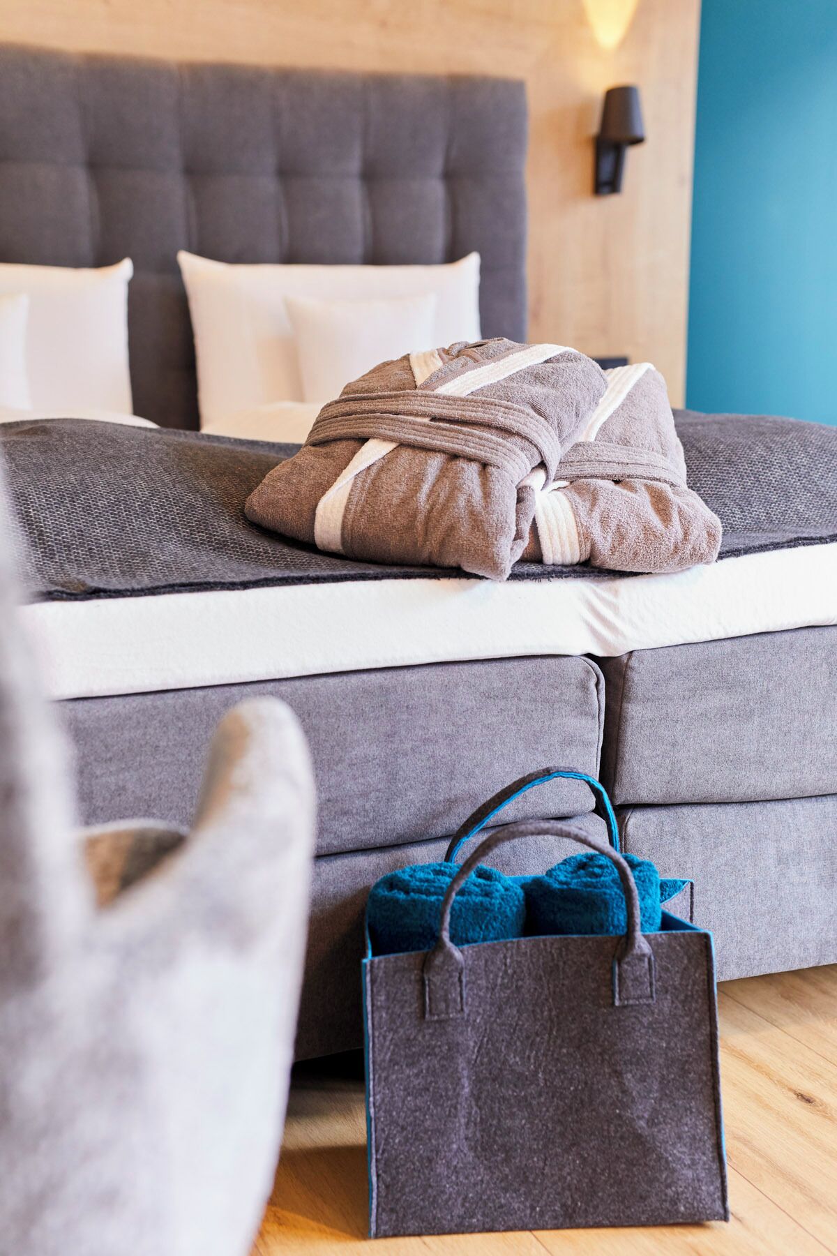Ein ordentlich bezogenes Bett mit einer grau-weißen Streifendecke, ein graues Sofa und eine blaue Tasche auf dem Boden.