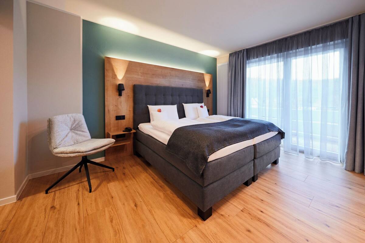 A modern hotel room with a large bed, a chair, and a view of the city through sheer curtains.
