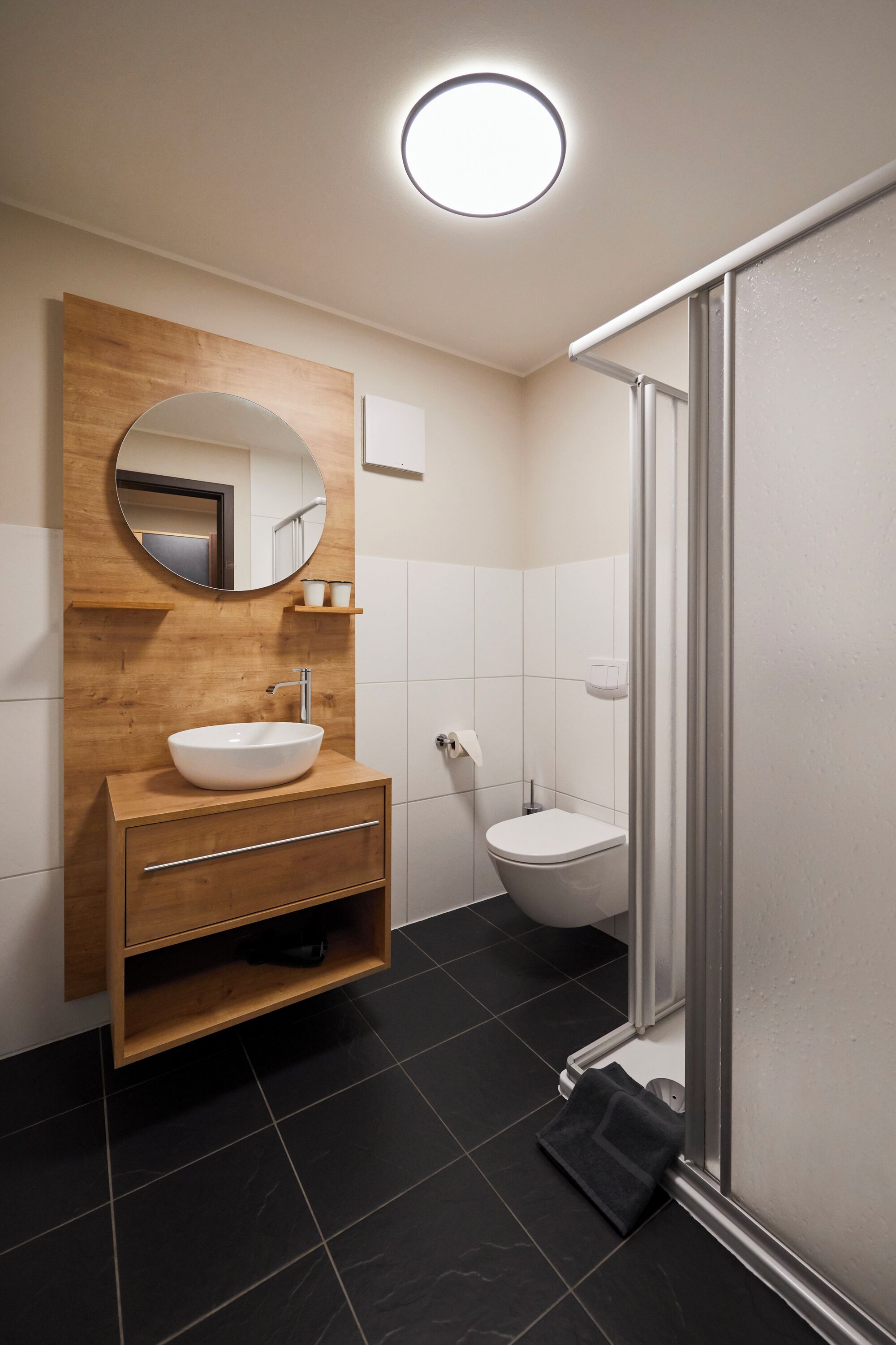 A modern bathroom with a wooden vanity, a round mirror, a white sink, a toilet, and a glass shower enclosure.