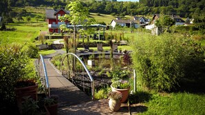 A rural landscape with a red house, a pond, and a pathway leading to a gazebo. - NaturPur Hotel Maarblick (Meerfeld)