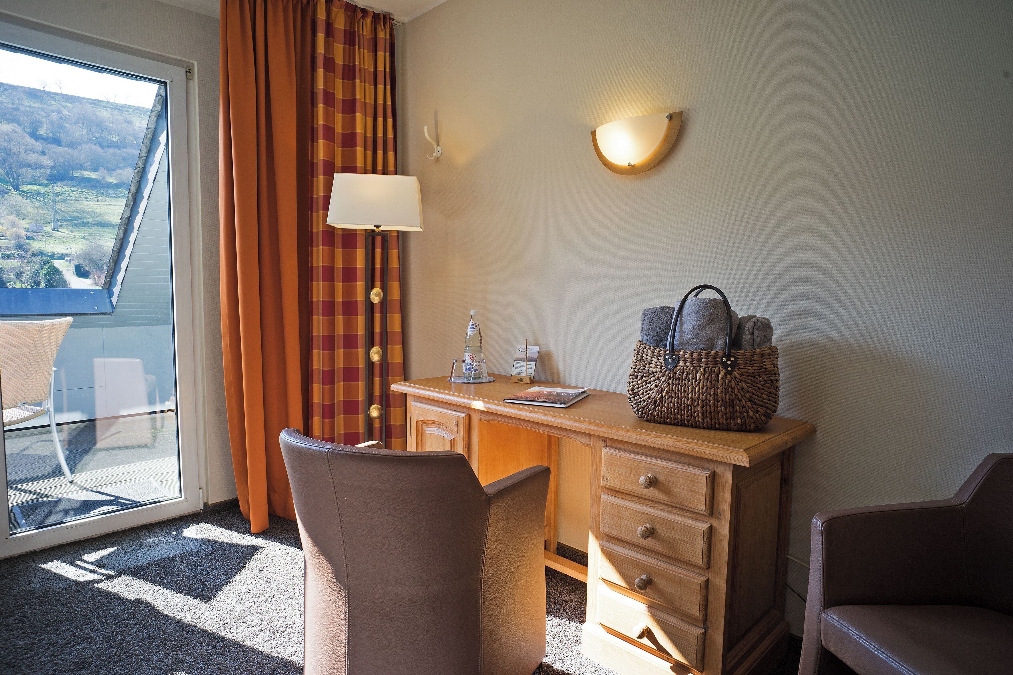 Habitación de hotel con escritorio, dos sillas, lámpara, cesta y vista al exterior.
