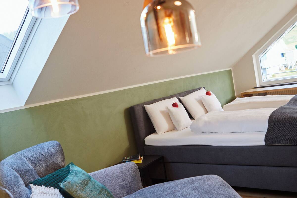 A modern room with a bed, a sofa, and a chair. There are pendant lights above the bed.