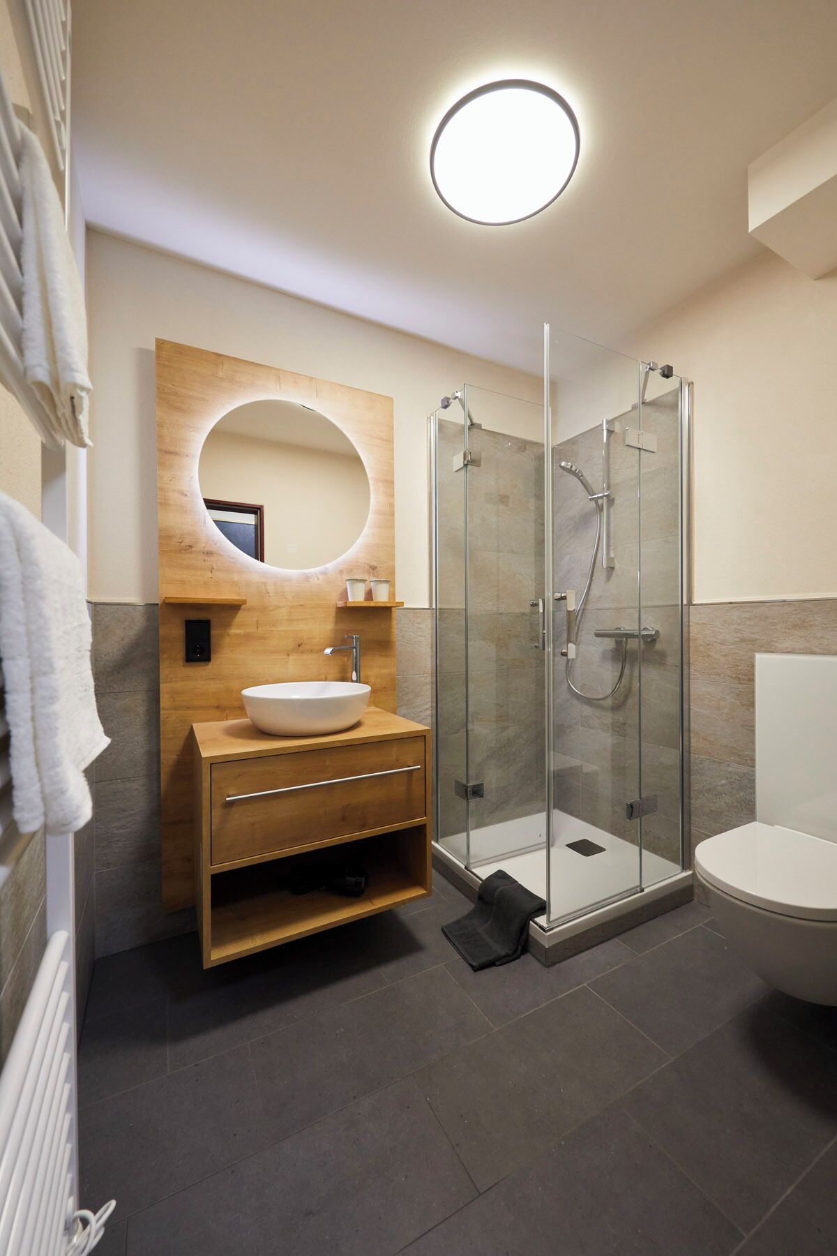 A modern bathroom with a wooden vanity, a round mirror, a glass-enclosed shower, and a toilet.