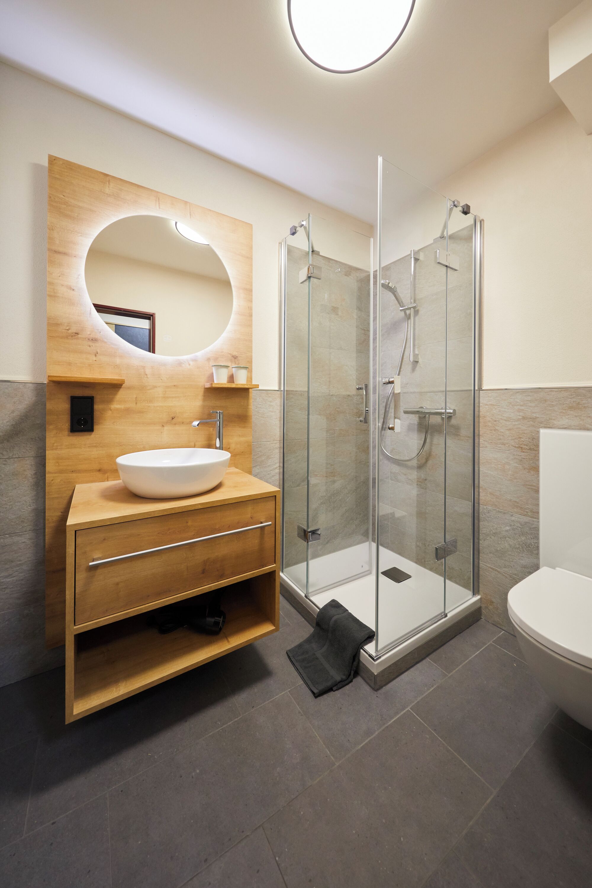 Un baño moderno con tocador de madera, un espejo grande, ducha con cerramiento de vidrio y un inodoro blanco.