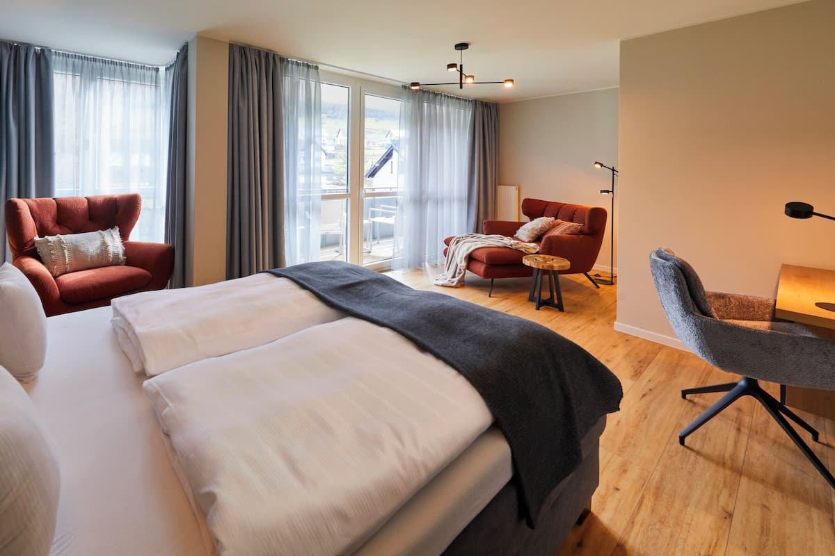 A modern hotel room with a large bed, two armchairs, a small table, and a view of the outdoors through large windows.
