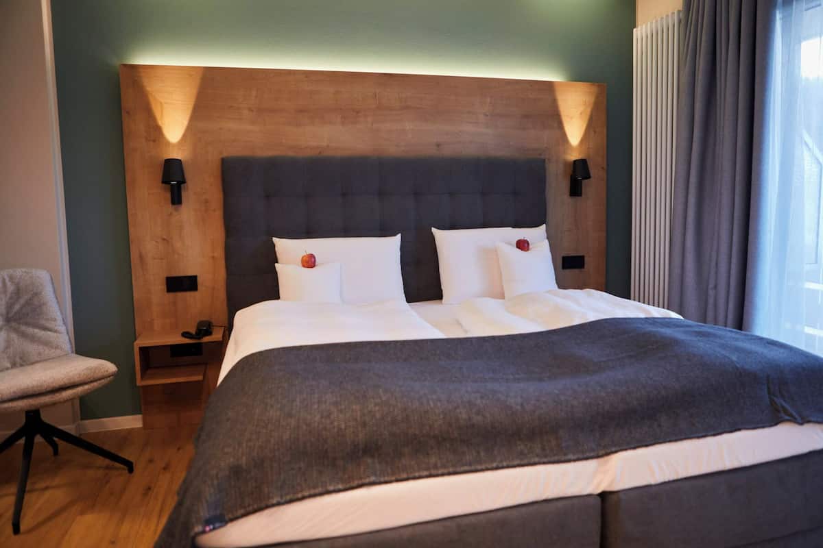 A modern hotel room with a large bed, two bedside tables, a chair, and a wooden headboard.