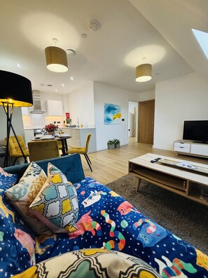 Una sala moderna con un sofá, una mesa de centro y un televisor. Se observa una cocina al fondo con gabinetes blancos y un comedor con sillas amarillas.