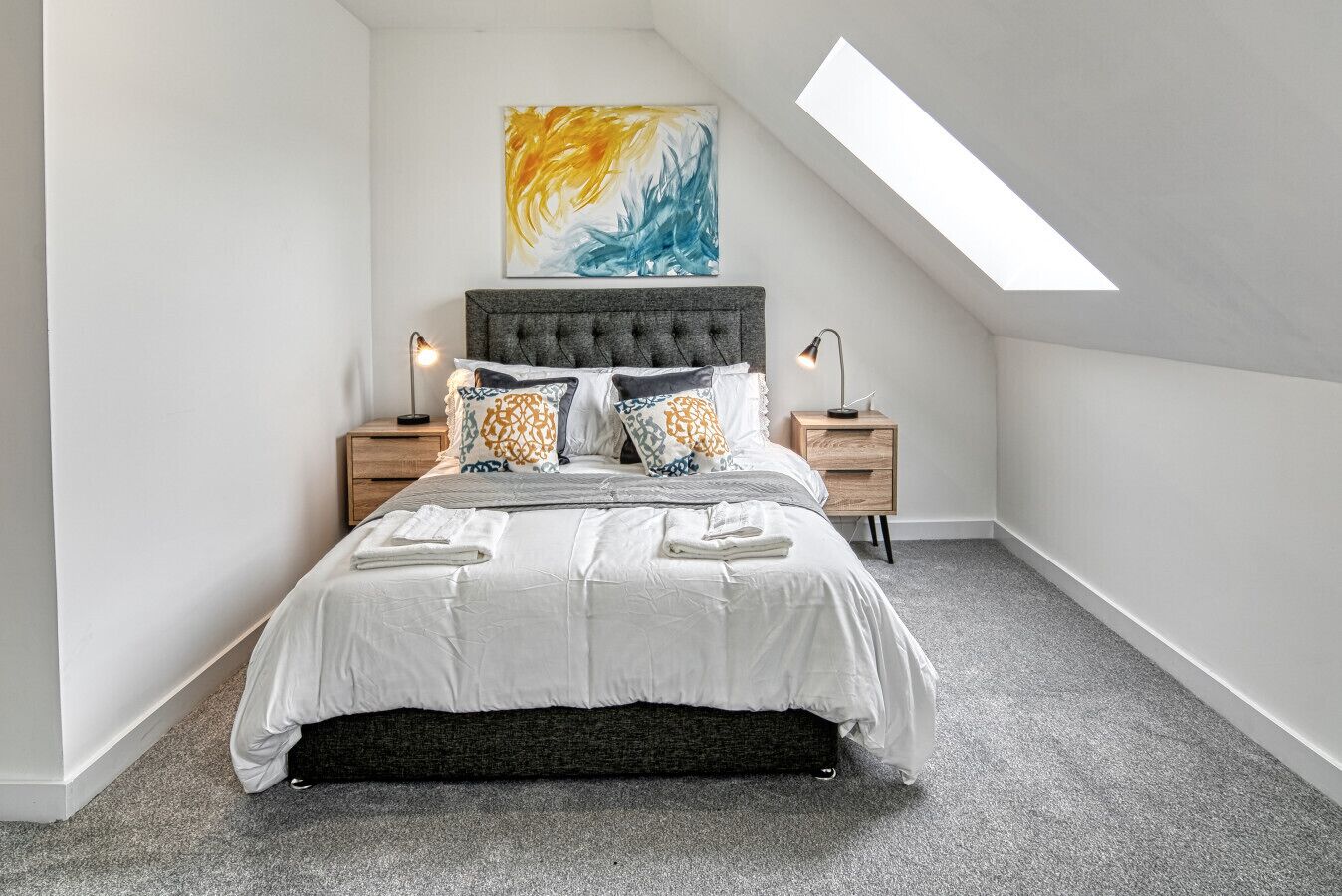 A bedroom with a sloped ceiling, a bed with white bedding, multiple pillows, two bedside tables with lamps, and a large abstract painting on the wall.