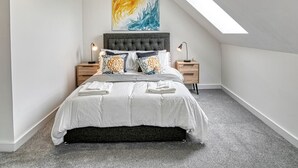 Une chambre avec un plafond en pente, un lit recouvert de linge de lit blanc, de nombreux oreillers, deux tables de chevet avec des lampes et un grand tableau abstrait accroché au mur.