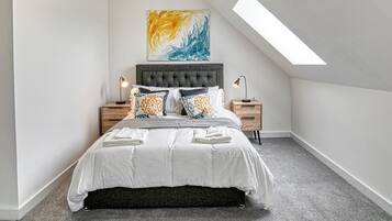 A bedroom with a sloped ceiling, a bed with white bedding, multiple pillows, two bedside tables with lamps, and a large abstract painting on the wall.