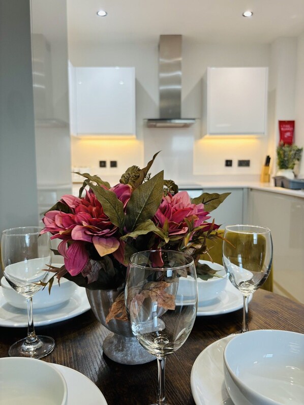Una mesa de comedor dispuesta con platos blancos, copas de vino transparentes y un centro de mesa de flores rosas y hojas secas.