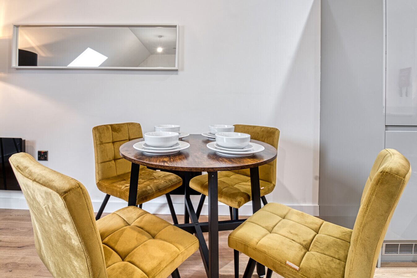A dining area with a round table and four mustard-colored chairs. There is a mirror on the wall above the table.