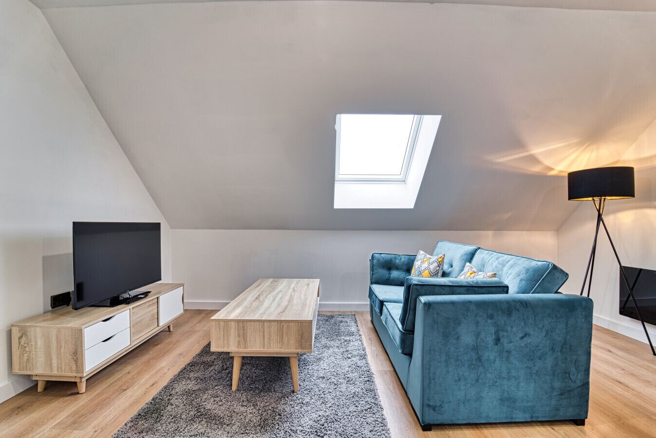 A modern loft apartment with a sloped ceiling, a skylight, a flat-screen TV, a wooden coffee table, a blue sofa, and a floor lamp.